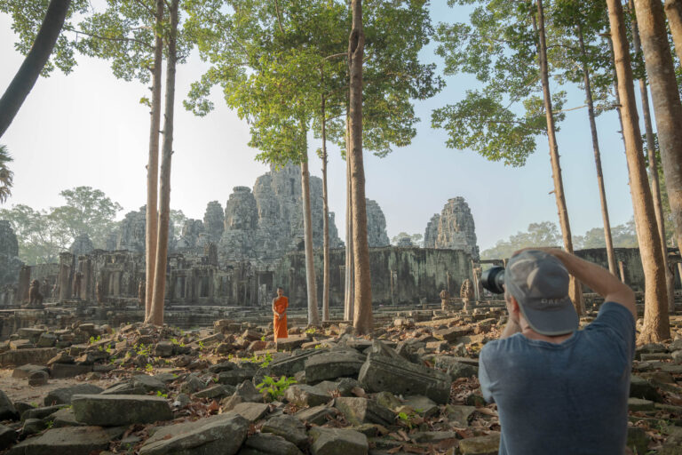 Photographer focusing camera lens on Bayon Temple stone face towers and a Buddhist monk in orange robes