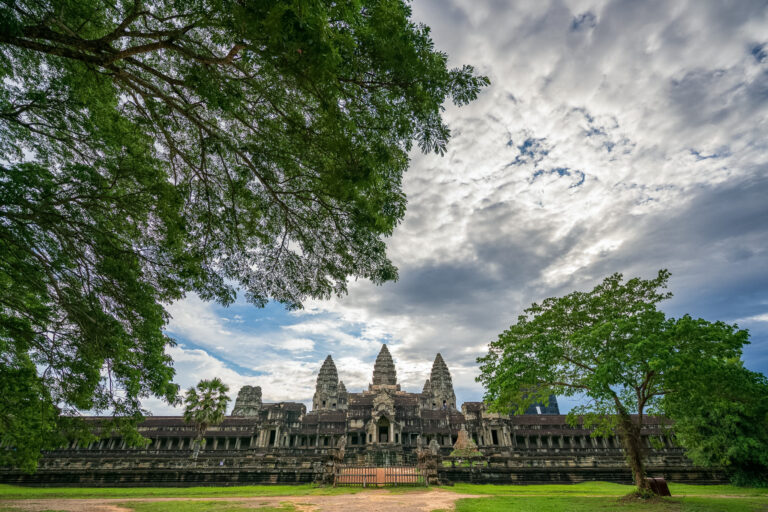 Quiet eastern view of Angkor Wat back entrance towers framed by large leafy green trees under dramatic clouds