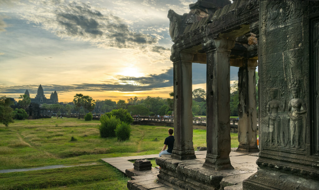 A visitor sitting on the stone wall of the outer West Gatehouse framed by pillars, watching the sun set over the causeway at Angkor Wat