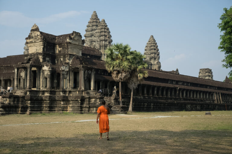 A monk in an orange robe walking across the grounds of Angkor Wat, with the temple's towers in the background.