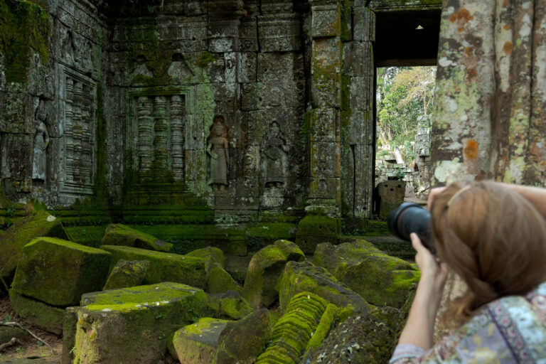Female photographer holding a camera up to capture moss-covered devata carvings at Ta Nei temple in Siem Reap